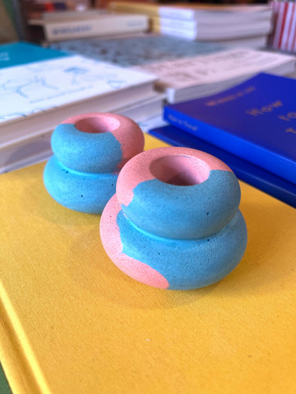 Two small donut-shaped ceramic objects in blue and pink, placed on a yellow surface with books in the background.