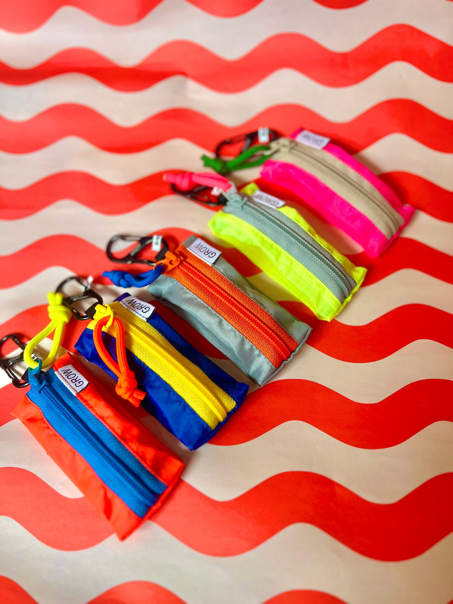Six small zippered pouches or keychain cases in assorted bright colors, arranged diagonally on a red and white wavy striped background. The pouches include blue, yellow, orange, red, green, and pink, each with a metal keychain clip.